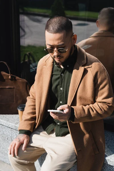 Trendy asian man using mobile phone while sitting on urban street — Stock Photo