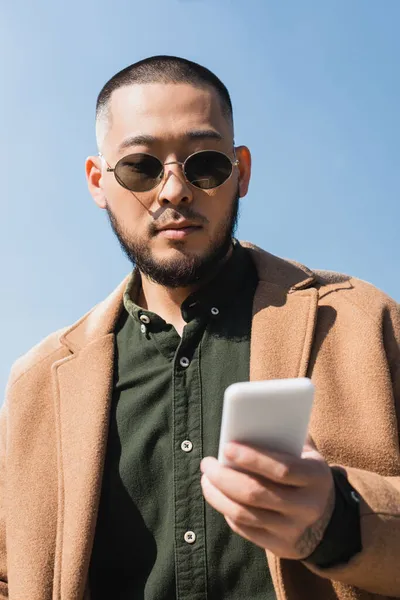 Low angle view of asian man in coat and sunglasses using blurred smartphone against blue sky — Stock Photo