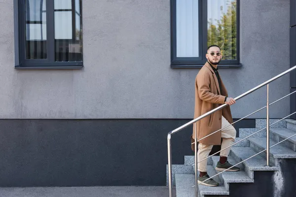 Full length view of trendy asian man walking upstairs near grey building — Fotografia de Stock