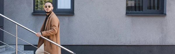 Hombre asiático en traje de otoño de moda y gafas de sol en las escaleras cerca de edificio gris, pancarta - foto de stock