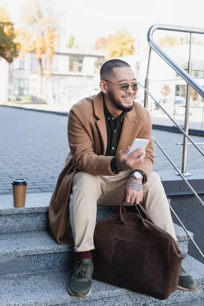 Glücklicher und stilvoller asiatischer Mann mit Handy sitzt auf einer Treppe in der Nähe von Pappbechern im Freien — Stockfoto