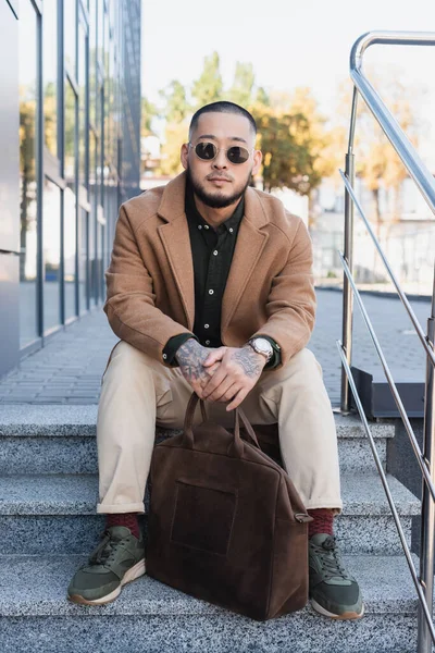 Stylish asian man in beige coat and sunglasses sitting on stairs with leather briefcase — Stock Photo