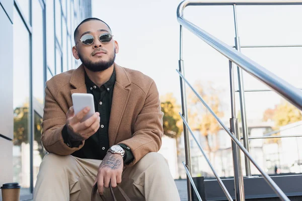 Trendiger asiatischer Mann in Mantel und Sonnenbrille sitzt mit Smartphone auf urbaner Straße — Stockfoto