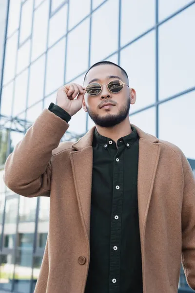 Bärtiger asiatischer Mann im trendigen Herbstmantel berührt Sonnenbrille auf urbaner Straße — Stockfoto