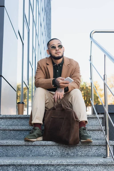 Bajo ángulo vista de asiático hombre en abrigo y gafas de sol sentado en escaleras con teléfono inteligente y maletín - foto de stock