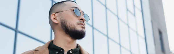 Bajo ángulo vista de asiático hombre en elegante gafas de sol mirando lejos cerca borrosa edificio con fachada de vidrio, bandera - foto de stock