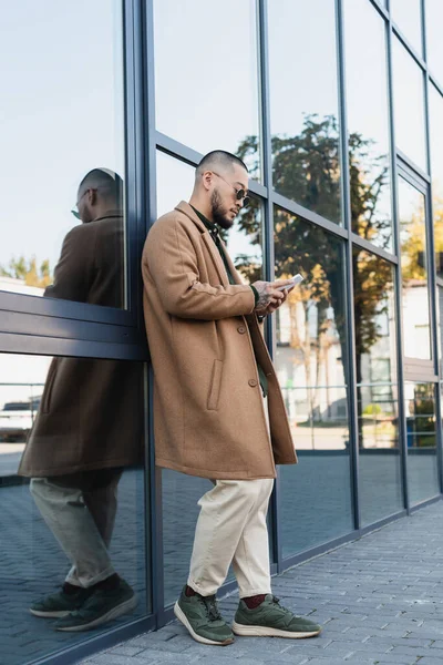 Повний вигляд на азіатського чоловіка в осінньому вбранні обміну повідомленнями на мобільному телефоні поблизу будівлі — стокове фото