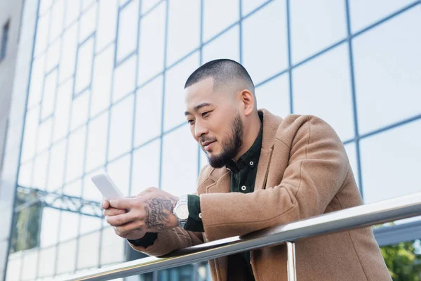 Tattooed asian man in beige coat messaging on smartphone near building with glass facade — Stock Photo