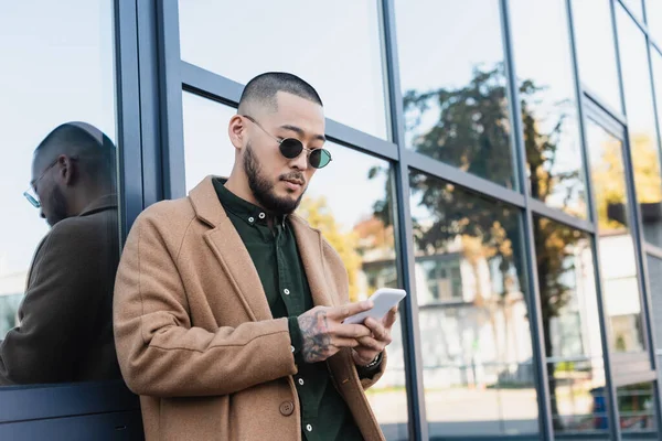 Азіатський чоловік в модних сонцезахисних окулярах обміну повідомленнями на смартфоні біля скляного фасаду будівлі — стокове фото