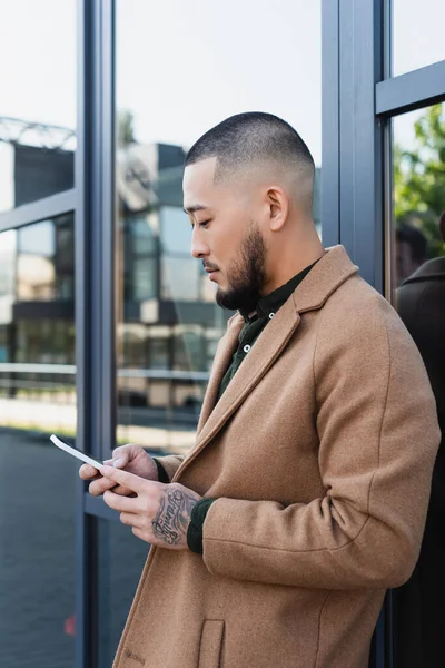 Asian man in autumn coat using mobile phone near glass facade of building — Stock Photo