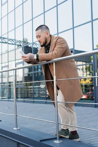 Asian man in beige coat leaning on metal fence while messaging on mobile phone outdoors — Stock Photo