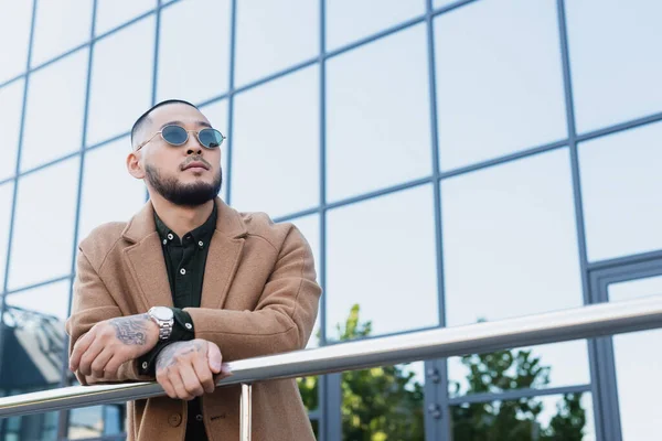 Tattooed asian man in beige coat leaning on metal fence near building with glass facade — Stock Photo