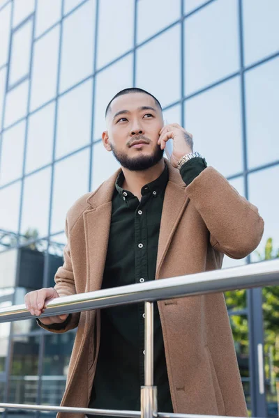 Asian man in autumn coat talking on smartphone near metal fence and building with glass facade — Stock Photo