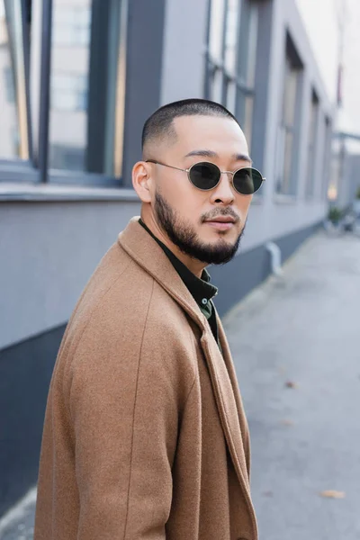 Bearded asian man in autumn coat and sunglasses looking at camera near blurred building — Stock Photo