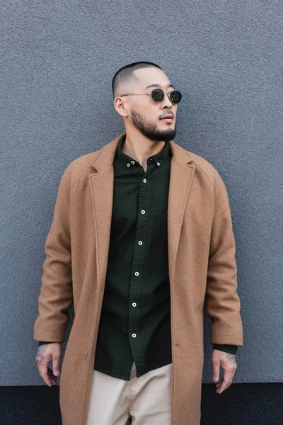 Barbudo asiático hombre en otoño abrigo y gafas de sol de pie cerca de gris pared — Stock Photo