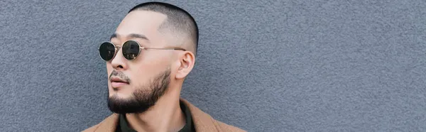 Bärtiger asiatischer Mann mit trendiger Sonnenbrille in der Nähe einer grauen Mauer im Freien, Banner — Stockfoto