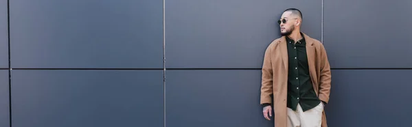 Стильний азіатський чоловік в бежевому пальто і сонцезахисних окулярах стоїть з рукою в кишені біля сірої стіни, банер — стокове фото