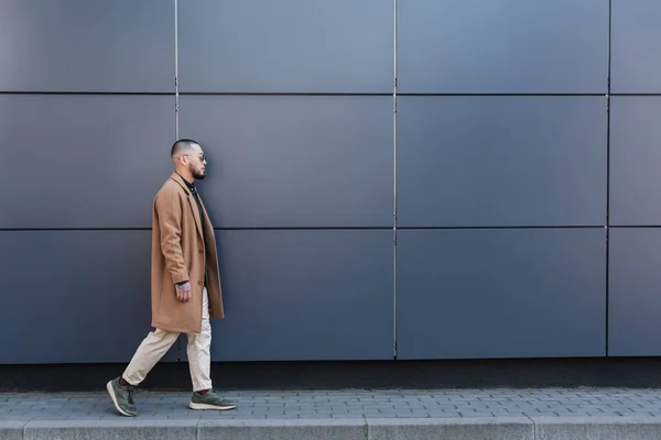 Вид сбоку на человека в бежевом пальто, идущего вдоль серой стены по городской улице — стоковое фото