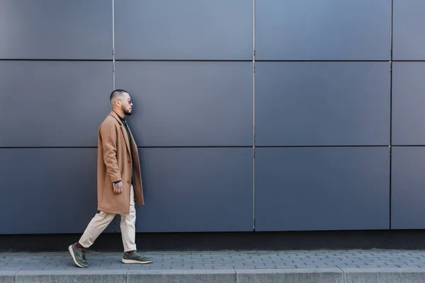 Side view of asian man in beige coat walking along grey wall on city street — Stock Photo
