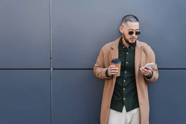 Trendy asian man with takeaway drink messaging on mobile phone near grey wall — Stock Photo