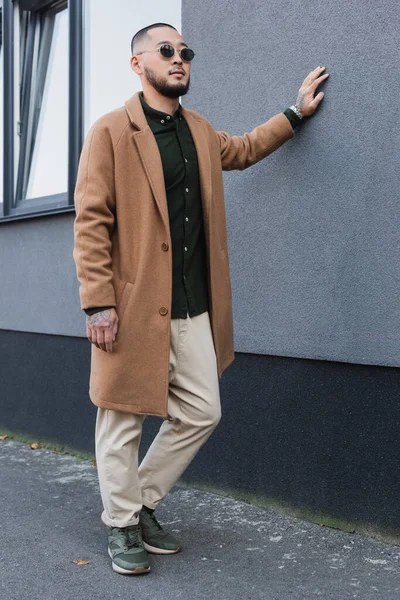 Asian man in trendy coat and sunglasses leaning on grey building on urban street — Stock Photo