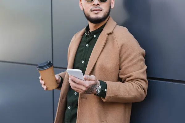 Cropped view of man in beige coat standing with coffee to go and smartphone near grey wall — Stock Photo