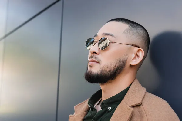 Asiático hombre en moderno gafas de sol de pie cerca gris pared al aire libre - foto de stock