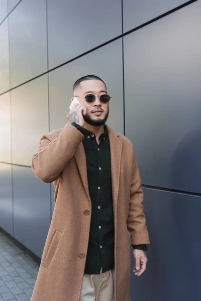 Asiatischer Mann mit Sonnenbrille und beigem Mantel telefoniert im Freien in der Nähe einer grauen Wand — Stockfoto