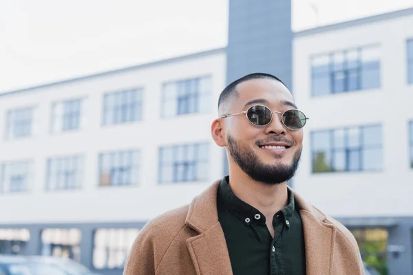 Alegre asiático hombre en elegante gafas de sol sonriendo cerca borrosa edificio en urbano calle - foto de stock