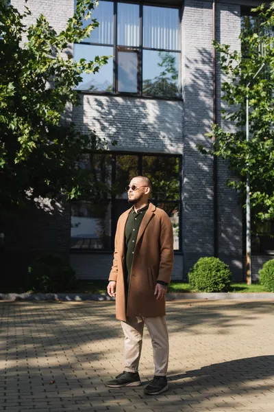 Full length view of asian man in stylish autumn outfit standing near building and trees on street — Stock Photo