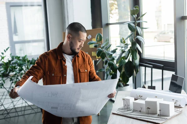 Arquitecto asiático mirando modelos de casa en el escritorio mientras sostiene el plano en la oficina - foto de stock