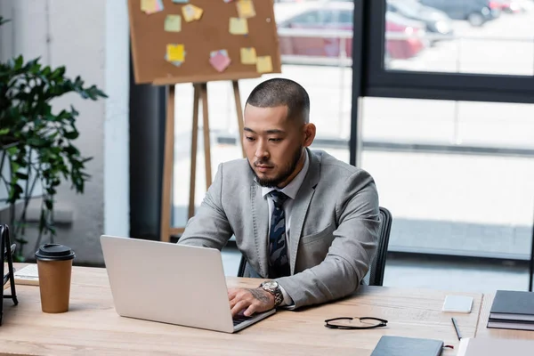 Asiatischer Geschäftsmann in offizieller Kleidung tippt auf Laptop in der Nähe von Imbiss-Drink auf dem Schreibtisch — Stockfoto