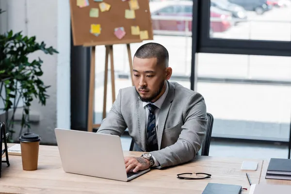 Asian businessman in formal wear typing on laptop near takeaway drink on desk — Stock Photo