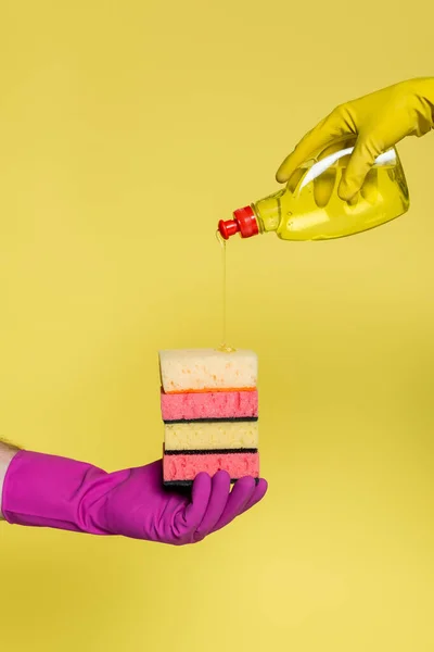 Visão recortada de pessoas em luvas de borracha segurando garrafa com detergente e pilha de esponjas isoladas em amarelo — Fotografia de Stock