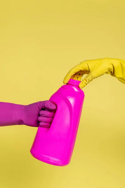 Vista recortada de las personas en guantes de goma sosteniendo botella de color rosa con detergente aislado en amarillo - foto de stock