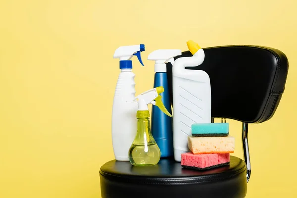 Flaschen mit Waschmittel und sauberen Schwämmen auf schwarzem Stuhl isoliert auf gelb — Stockfoto