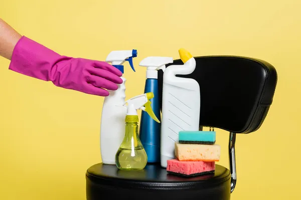 Обрезанный вид очистителя достигая бутылку с моющим средством рядом с чистыми губки на черном кресле изолированы на желтый — стоковое фото