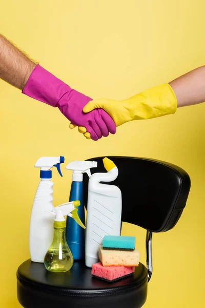 Abgeschnittene Ansicht von Reinigungskräften beim Händeschütteln in der Nähe von Flaschen mit Waschmittel und sauberen Schwämmen auf Stuhl isoliert auf gelb — Stockfoto