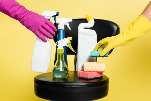 Cropped view of cleaners reaching bottle with detergent and sponge on black chair isolated on yellow — Stock Photo