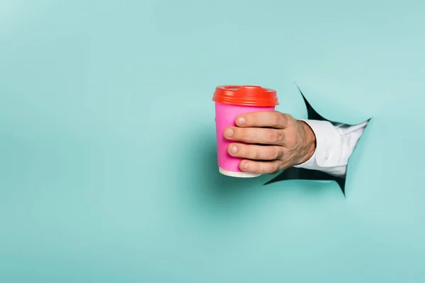 Cropped view of man holding coffee to go through hole in paper wall on blue — Stock Photo