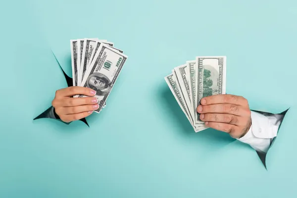 Cropped view of man and woman holding dollar banknotes through holes in paper wall on blue — Stock Photo