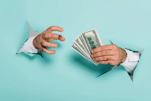 Cropped view of man holding dollar banknotes through holes in paper wall on blue — Stock Photo