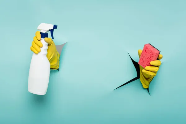 Partial view of cleaner in rubber gloves holding sponge and spray bottle through holes in paper wall on blue — Stock Photo