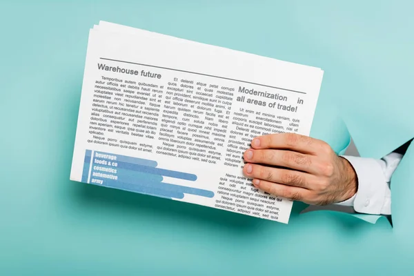 Cropped view of man holding newspaper through hole in paper wall on blue — Stock Photo