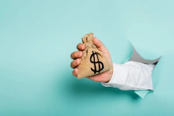 Cropped view of man holding money bag with dollar sign through hole in paper wall on blue — Stock Photo