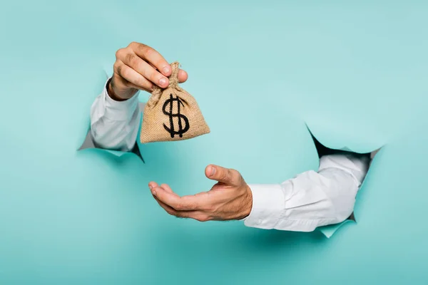 Partial view of man holding money bag with dollar sign through holes in paper wall on blue — Stock Photo