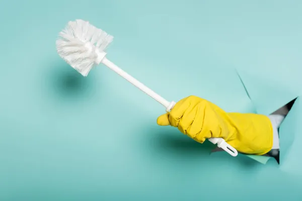 Vue partielle du nettoyant dans le gant en caoutchouc tenant la brosse de toilette à travers le trou dans le mur de papier sur bleu — Photo de stock