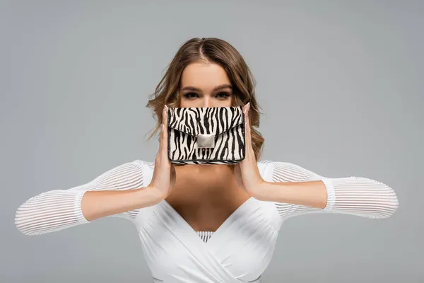 Curly young woman in white outfit covering face while holding purse with animal print isolated on grey — Stock Photo