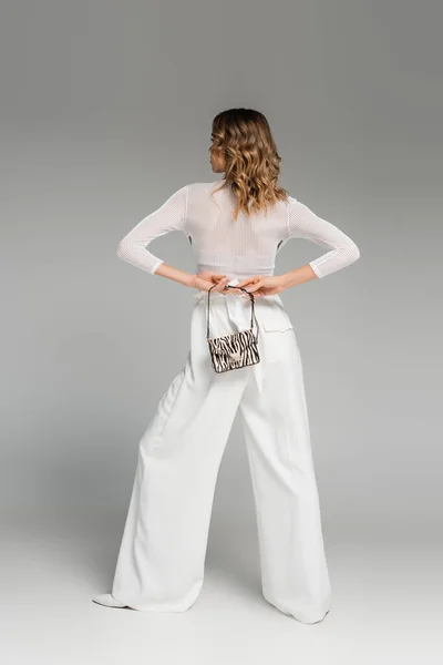 Full length of curly woman in white outfit holding purse with animal print behind back on grey — Stock Photo