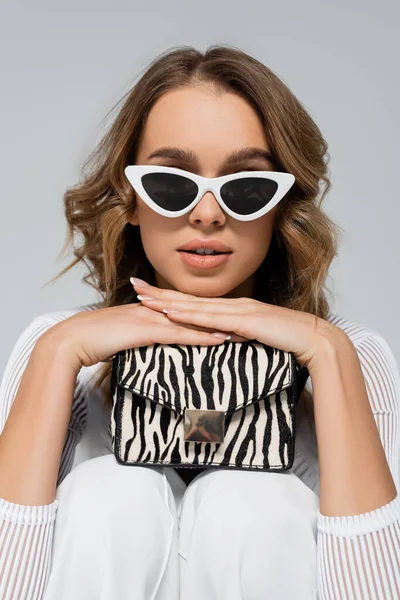 Curly woman in sunglasses holding purse with animal print isolated on grey — Stock Photo